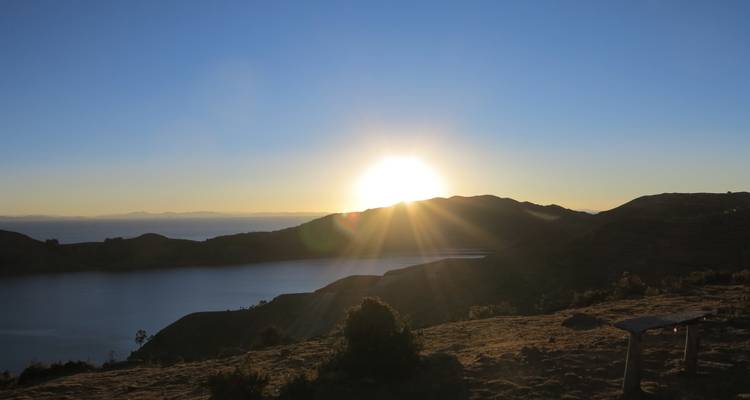 Amanecer sobre un lago tranquilo de gran altitud con cerros silueteados y rayos de sol radiantes.