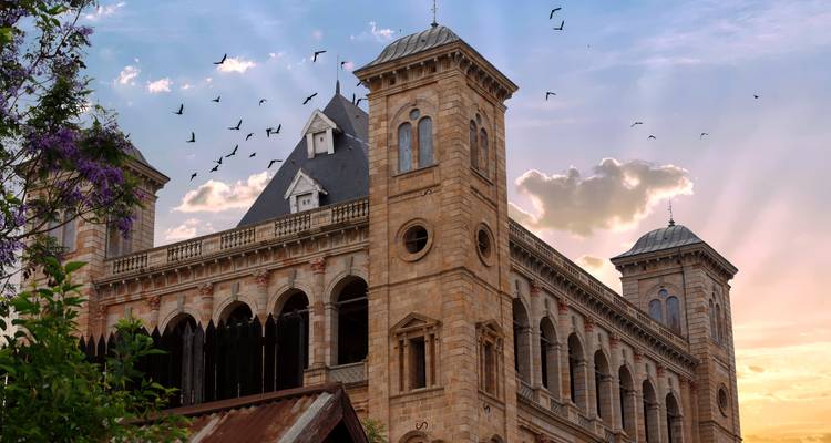 Palais Royal Historique d'Antananarivo avec des arches en pierre et des silhouettes d'oiseaux se détachant contre un ciel de coucher de soleil aux tons pastel.