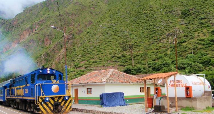 Locomotora azul del tren PeruRail pasando por una pequeña estación rural contra las laderas de montaña verde.