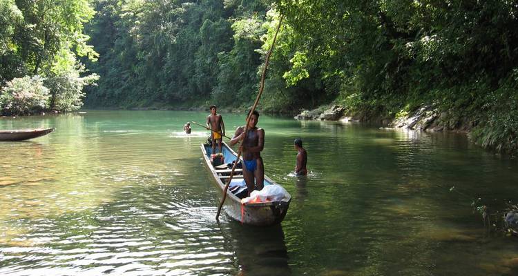 Indigene Maenner, die ein schmales Einbaum-Kanu durch einen ruhigen Dschungelfluss paddeln, waehrend sich andere in der Naehe baden.
