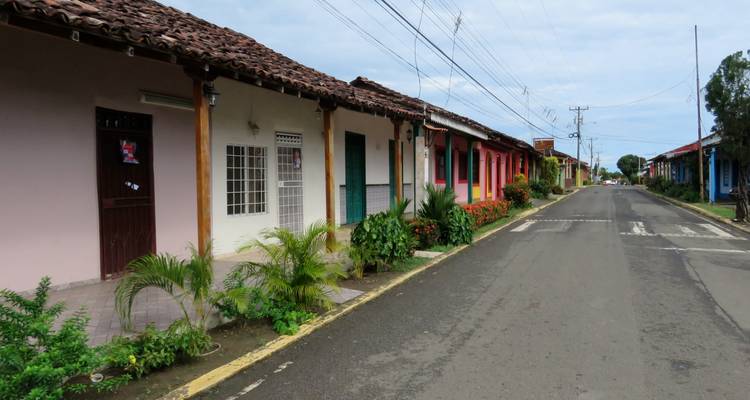 Calle tranquila de un pueblo bordeada de casas de una sola planta de colores vibrantes y plantas tropicales.