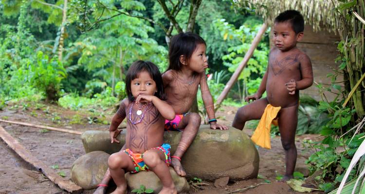 Tres niños indígenas con pintura corporal juegan en grandes rocas en un entorno de selva exuberante.