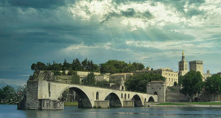 Historische stenen brug die een rivier overspant met het Pauspaleis van Avignon onder een dramatische hemel.