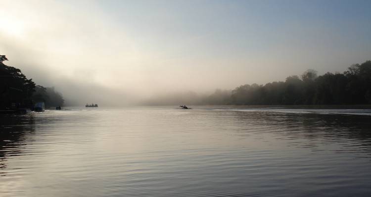 Nebeliger tropischer Fluss in der Morgenröte mit kleinen Booten, die kaum durch weichen Morgennebel sichtbar sind.
