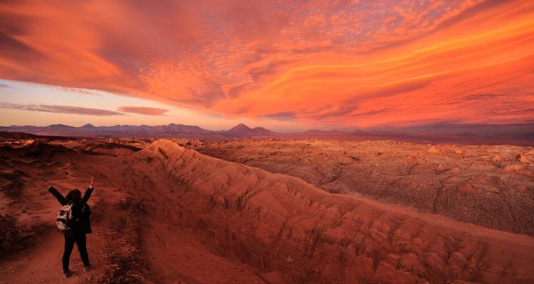 Voyageur les bras levés surplombant une vallée désertique rouge ardente sous un ciel de coucher de soleil épique à Atacama.