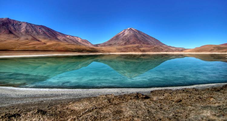 Le Laguna Verde aux eaux turquoise cristallines reflétant parfaitement un pic volcanique conique sous un ciel bleu.