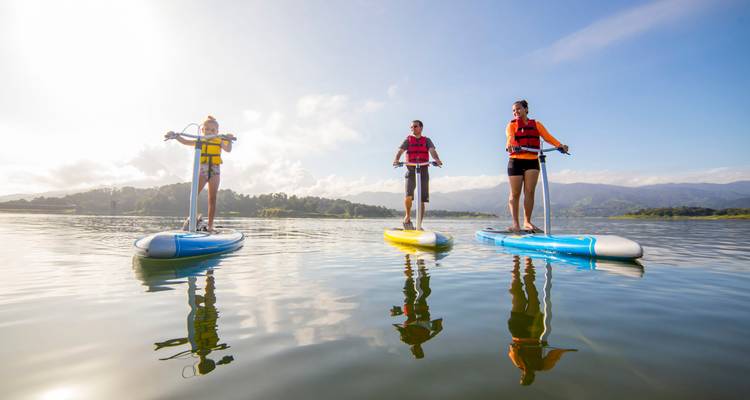 Tres personas practicando stand-up paddle en aguas tranquilas de un lago con montañas al fondo