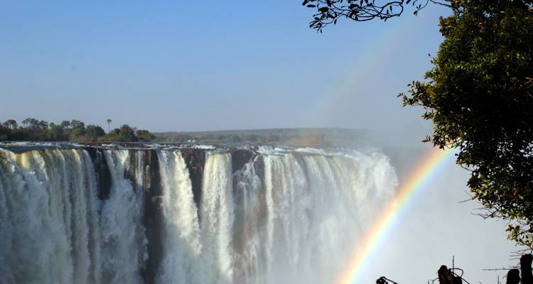 De bulderande Victoria Falls stuurt waterdruppels omhoog, wat een levendig regenboogje vormt tegen een heldere lucht.