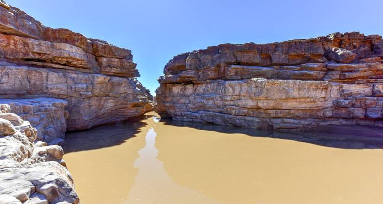 Rotswanden van een rotsachtige canyon weerspiegeld in een smalle modderige rivier onder een heldere blauwe hemel.