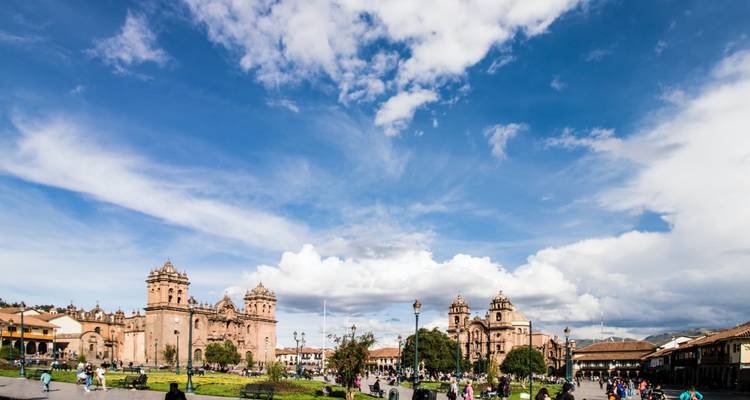 Panoramablick auf Cuscos Plaza de Armas mit doppelten Kathedralstürmen, Kolonialgebäuden und lebhaften Menschenmengen unter einem weiten blauen Himmel mit dramatischen Wolkenformationen.