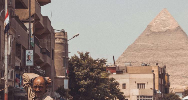 Une scène animée d'une rue du Caire avec des immeubles d'habitation au premier plan et la Grande Pyramide s'élevant à l'arrière-plan.