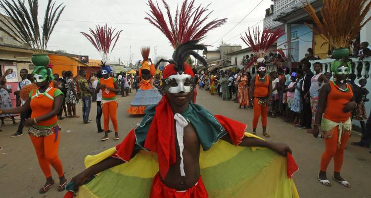 Desfile callejero con performers enmascarados en trajes naranja brillante y tocados con plumas rodeados de espectadores