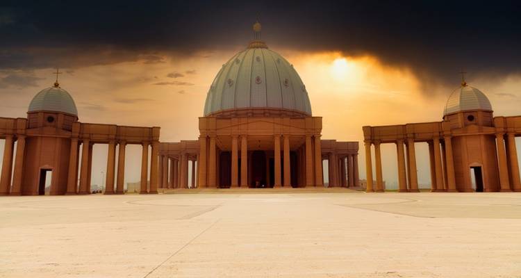 Gran basílica con una cúpula enorme y columnatas bajo unas nubes de atardecer dramáticas en una vasta plaza de mármol