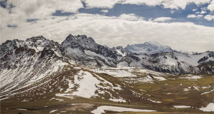 Des pics andins enneigés dominant un plateau d'altitude avec des taches de neige et des randonneurs en contrebas.