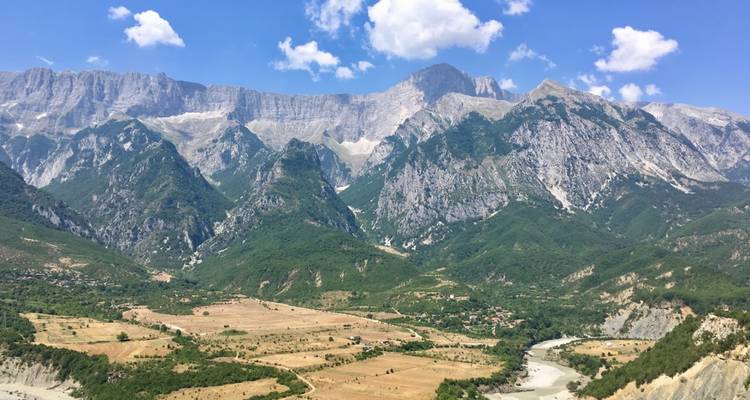 Des pics alpins spectaculaires dominant une vallée fertile et une rivière sinueuse sous un ciel bleu avec des nuages.