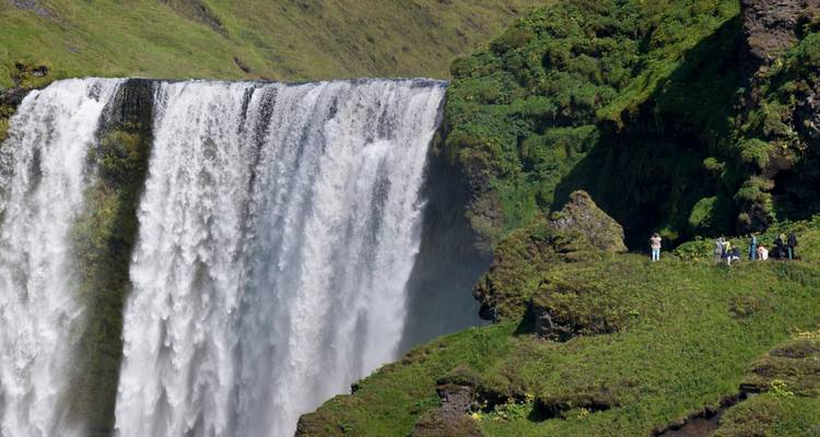 La puissante cascade de Skógafoss dévale les falaises verdoyantes aux côtés de minuscules touristes.