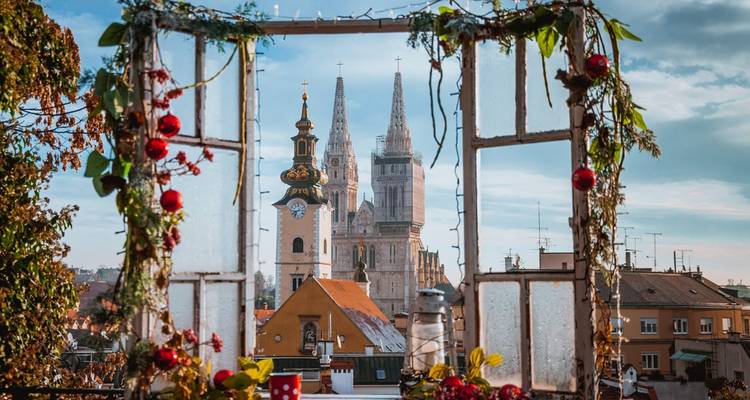 Vue de la Cathédrale de Zagreb encadrée par une fenêtre rustique décorée de fleurs et d'ornements.