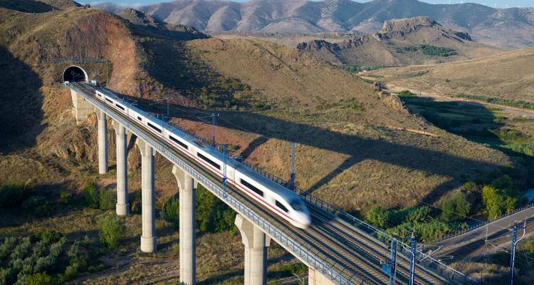 Train blanc à grande vitesse traversant un long viaduc qui émerge d'un tunnel dans les collines arides espagnoles.