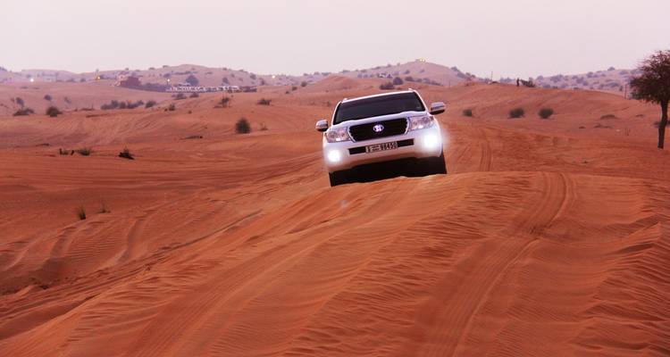 Un vehículo 4x4 sube la cresta de una duna de arena roja en el desierto al atardecer con los faros encendidos.