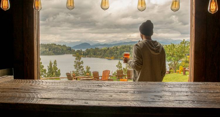 Person, die an einem rustikalen Barfenster mit Blick auf einen See und Berge an einem bewölkten Tag ein Getränk genießt.