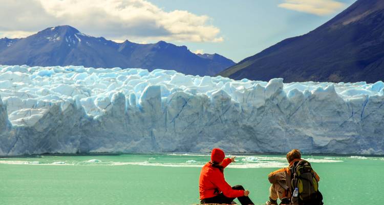 Zwei Wanderer ruhen sich am türkisfarbenen Wasser aus und blicken auf den aufragenden Perito-Moreno-Gletscher.
