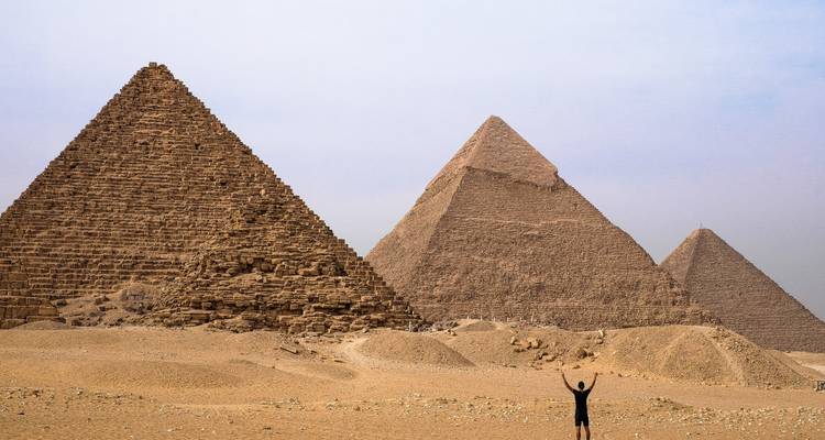 Un voyageur lève les bras devant les trois grandes pyramides sur le plateau désertique de Giza sous un ciel pâle