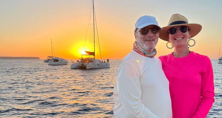 Couple de personnes âgées souriantes posant sur un bateau en mer lors d'un coucher de soleil éclatant avec des catamarans en arrière-plan.