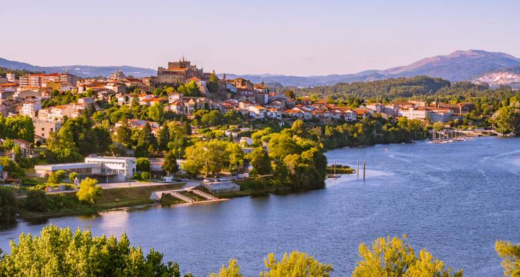 Ville portugaise au bord du fleuve couronnée par une église au sommet de la colline et un château sous un ciel du soir dégagé.