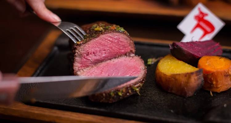Nahaufnahme eines saftigen, medium-rare gegarten Steaks, das auf einem heißen Steinplatte mit Gemüse geschnitten wird