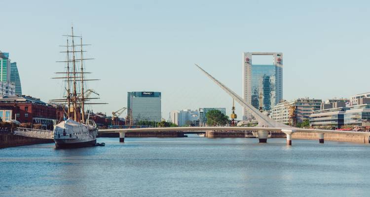 Moderne Skyline und die elegante Puente de la Mujer Brücke, die ein ruhiges Ufergebiet bei hellem Tageslicht überspan­nt.