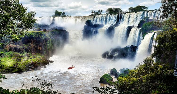Breites Panorama der Iguazu-Fälle mit einem kleinen Touristenboot, das die Gischt darunter trotzt.