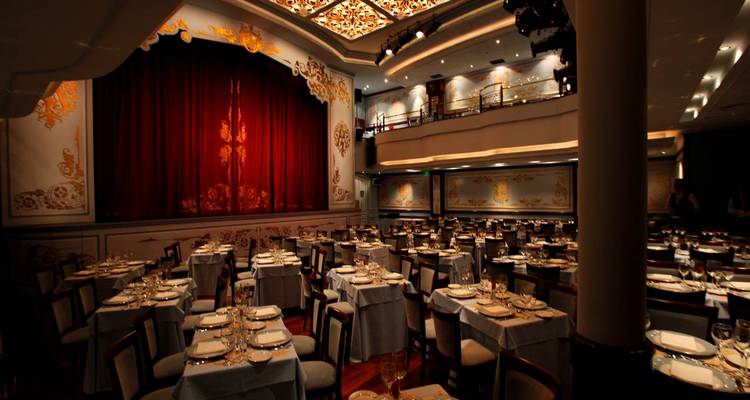 Elegantes Dinner-Theater mit reservierten Tischen, die auf eine mit rotem Vorhang verhängte Bühne ausgerichtet sind.