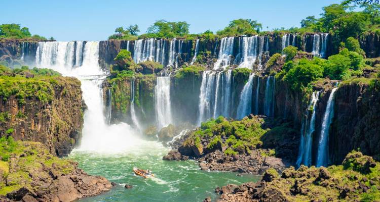 Panoramischer Tagesansicht mehrerer Wasserfälle der Iguazu-Fälle inmitten üppigen grünen Dschungels mit Touristenboot darunter.