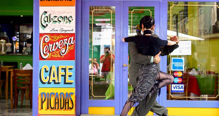 Ein Paar führt einen energievollen Tango vor einem farbenfrohen Café auf, das mit Vintage-Schildern und leuchtend violetten Türen geschmückt ist.