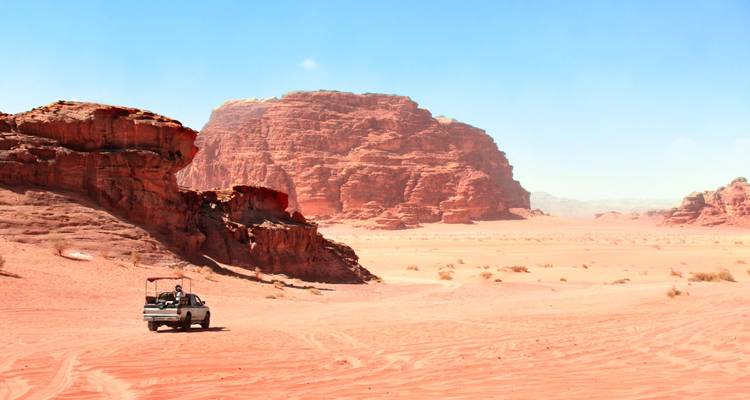 Pickup truck met toeristen rijdt over een uitgestrekte rode woestijnvlakte omringd door zandstenen mesa's