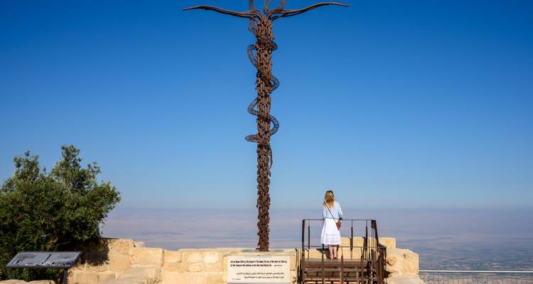 Een vrouw in het wit kijkt naar een hoog bronzen kronkelend kruis monument bovenop een stenen terras met uitgestrekte woestijndalvistas onder een heldere blauwe lucht.