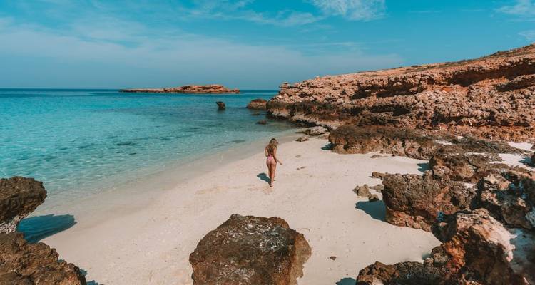 Une femme se promène le long d'une plage isolée aux sables blancs avec une eau turquoise et des promontoires rocheux sous un ciel bleu brillant.
