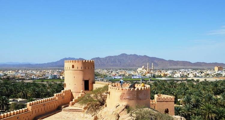 Les créneaux jaune-brun du fort de Nizwa s'élèvent au-dessus des palmeraies avec les montagnes et la ligne d'horizon de la ville en arrière-plan.