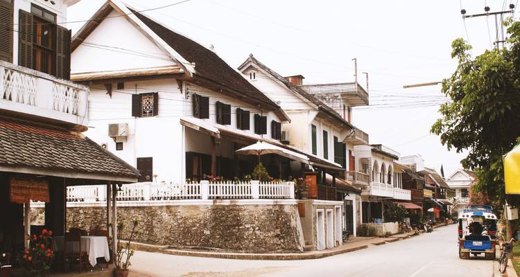 Rustige koloniale straat met witgeverfte huizen met luiken en een tuk-tuk geparkeerd onder bewolkte lucht in Luang Prabang.