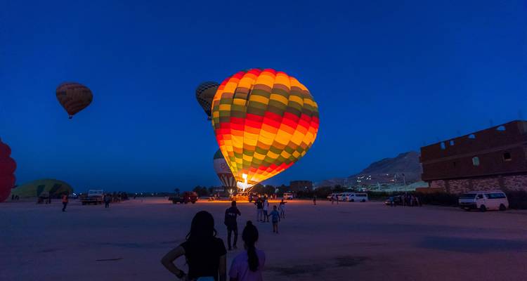 Un ballon à air chaud aux couleurs vives se gonfle sur une plaine désertique avant le lever du soleil tandis que des spectateurs se rassemblent.