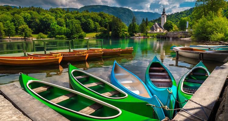 Rangée de canoës colorés amarrés sur un lac alpin serein avec un clocher d'église et des collines boisées au-delà.
