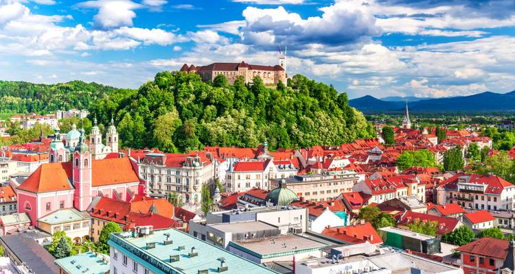 Vue panoramique sur Ljubljana avec le château sur une colline boisée et les rangées de bâtiments aux toits rouges en contrebas, encadrées par les montagnes et les nuages épars.