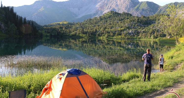 Oranges Campingzelt auf grünem Seeufer mit zwei Campern, die einen ruhigen Alpensee und die umliegenden grünen Berge bei Sonnenuntergang betrachten.