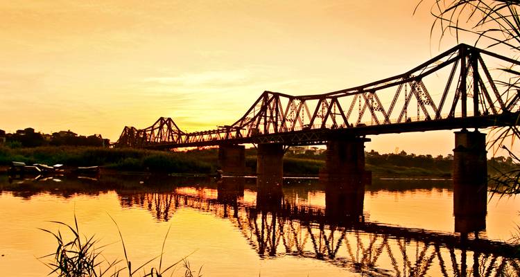 Die historische Long-Bien-Brücke überspannt einen reflektierenden Fluss unter einem orangefarbenen Himmel bei Sonnenuntergang in Hanoi.