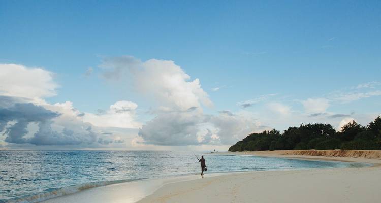 Pêcheur solitaire lançant sa ligne sur une plage de sable blanc immaculée sous un vaste ciel bleu