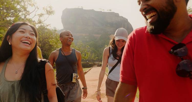 Cuatro viajeros felices caminando por un sendero con la enorme Roca de Sigiriya elevándose detrás de ellos bajo la luz brillante del sol.