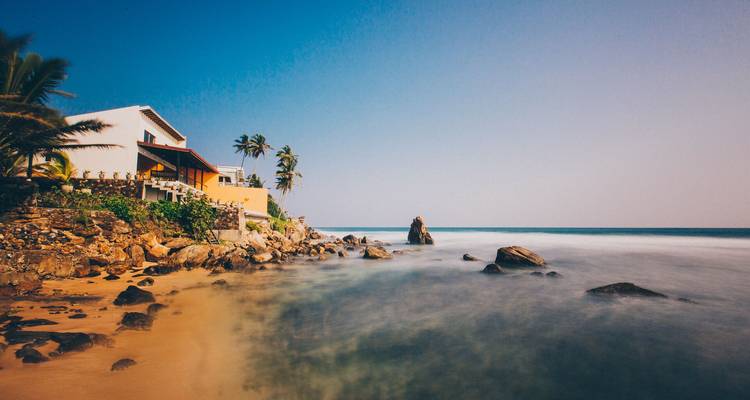 Escena de playa tropical con una villa moderna, palmeras y una costa rocosa junto a un océano turquesa tranquilo bajo un cielo azul despejado.