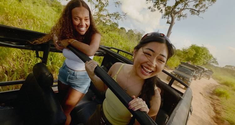 Dos mujeres jóvenes riéndose mientras están de pie en un jeep safari descapotable conduciendo por un camino polvoriento bordeado de pasto alto.