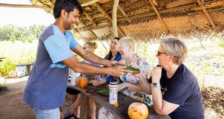 Lokale gastheer serveert voedsel verpakt in bananenbladen aan lachende reizigers onder een rieten afdak.