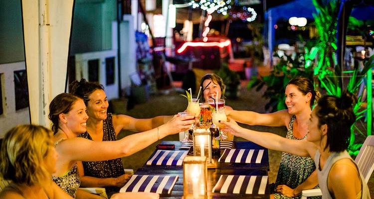 Groep vrienden klinken met tropische cocktails op een kaarsverlichtte tafel aan zee in de avond.